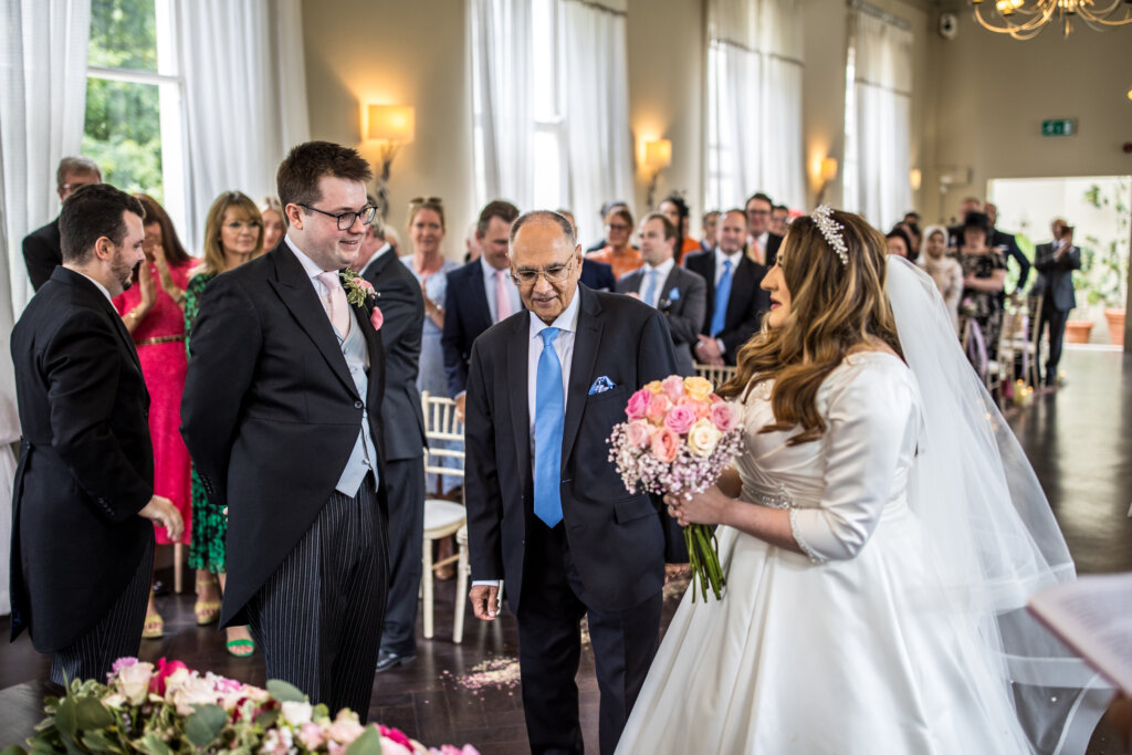 Groom seeing bride walk down the aisle with her father during ceremony in Mulberry Room at Morden Hall