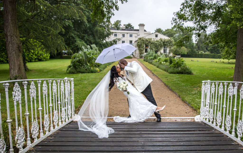 Bride and groom holding a white umbrella on the bridge with Morden Hall in the background