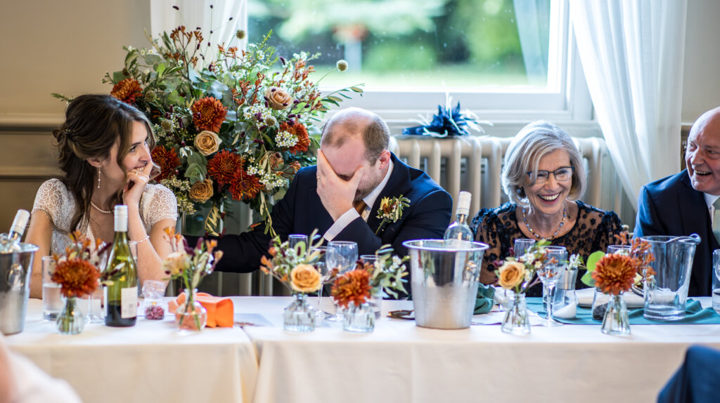 Groom covering his face in embarrassment during best man speech in Mulberry Room at Morden Hall
