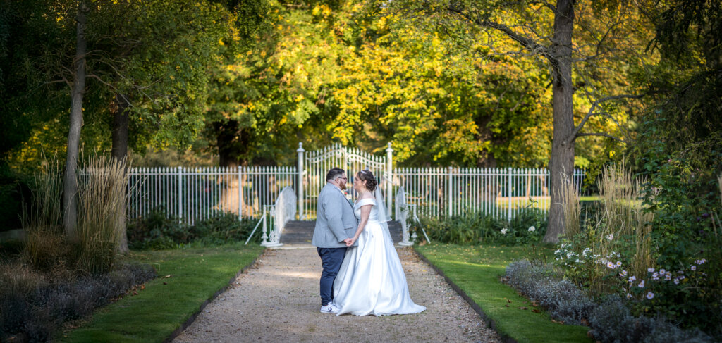 Bride and groom kissing on path at twilight with bridge and sunlit trees at Morden Hall