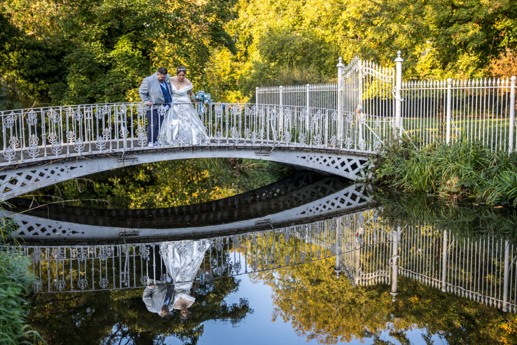Bride and groom on footbridge looking into River Wandle during golden hour sunset at Morden Hall