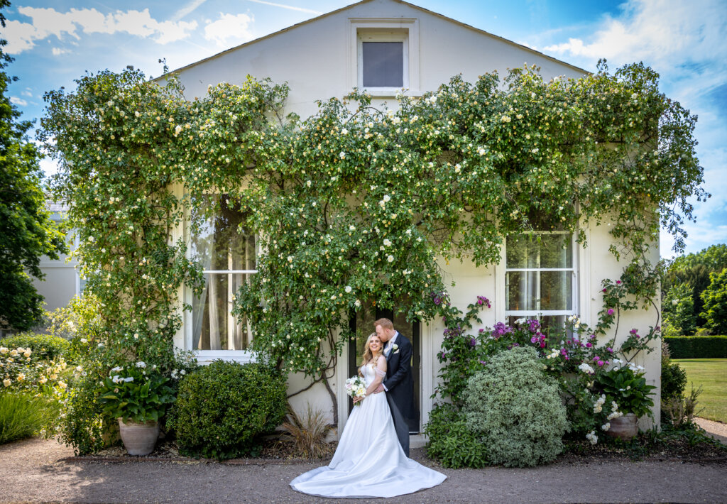 Bride and groom embracing outside the Willow Room doorway at Morden Hall framed by climbing roses