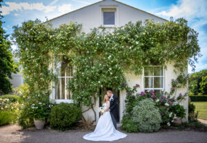 Bride and groom embracing outside the Willow Room doorway at Morden Hall framed by climbing roses