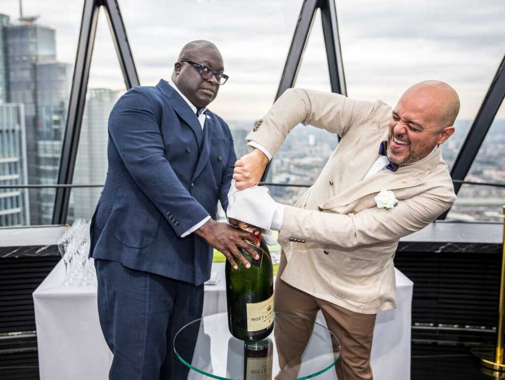 Two grooms opening a large bottle of Champagne.
